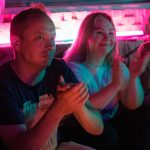 A lively group of adults enjoying a vibrant indoor event, illuminated by neon lights.